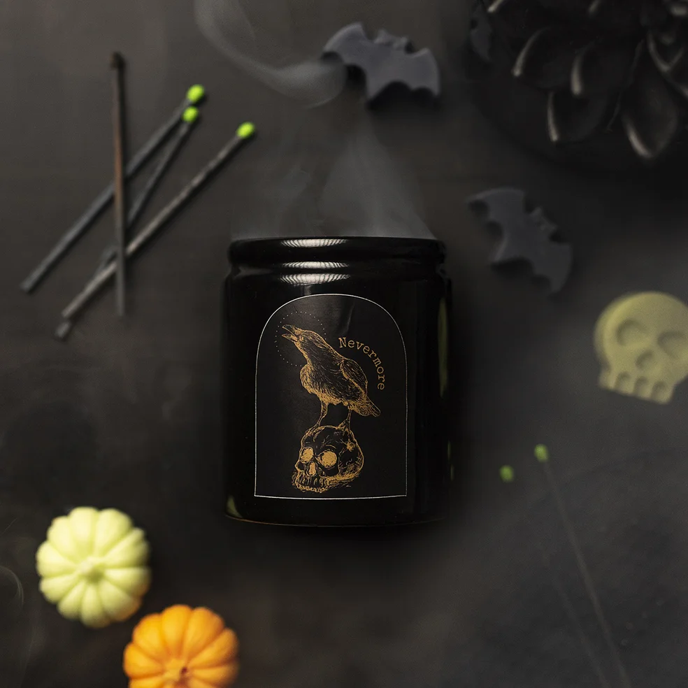 A black ceramic candle jar with a label showing the outline of a gold raven on a skull. 