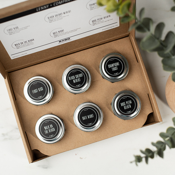 An overhead view of a candle sampler set: a brown cardboard box holds six candles poured in silver metal tins