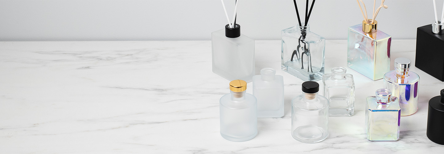 A variety of frosted, prism, and matte black reed diffuser bottles with reeds on display on white marble countertop.