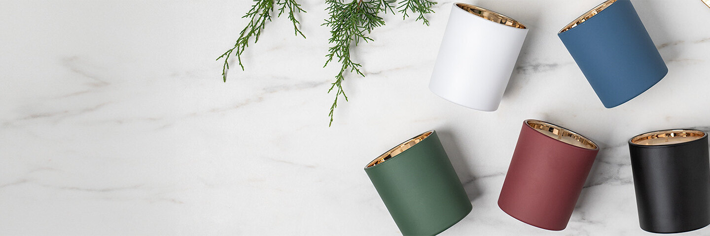 white, blue, red, green, and black tumbler jars with a warm gold interior displayed on a white marble countertop with evergreen and gold leaves.