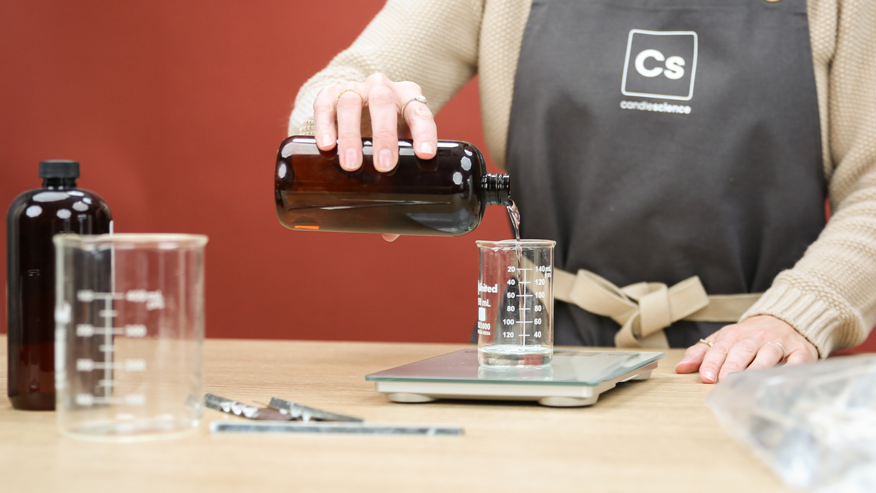 A light-skinned person wearing a grey CandleScience apron pours fragrance oil into a glass beaker on a scale