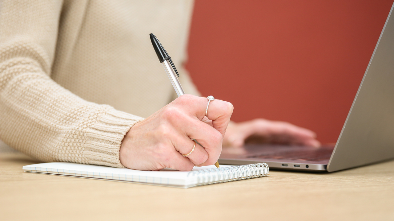 A pair of light-skinned hands writes notes on a pad of paper next to an open laptop