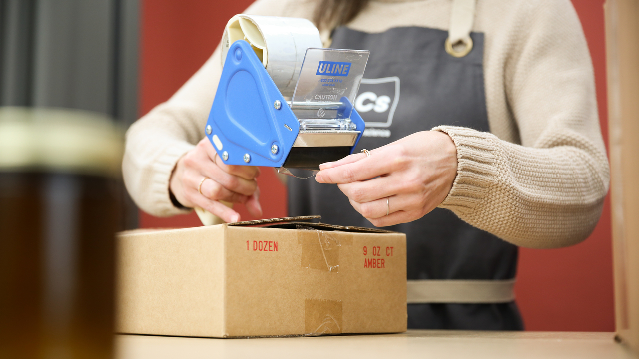 A light-skinned person tapes closed a cardboard box of handmade candles