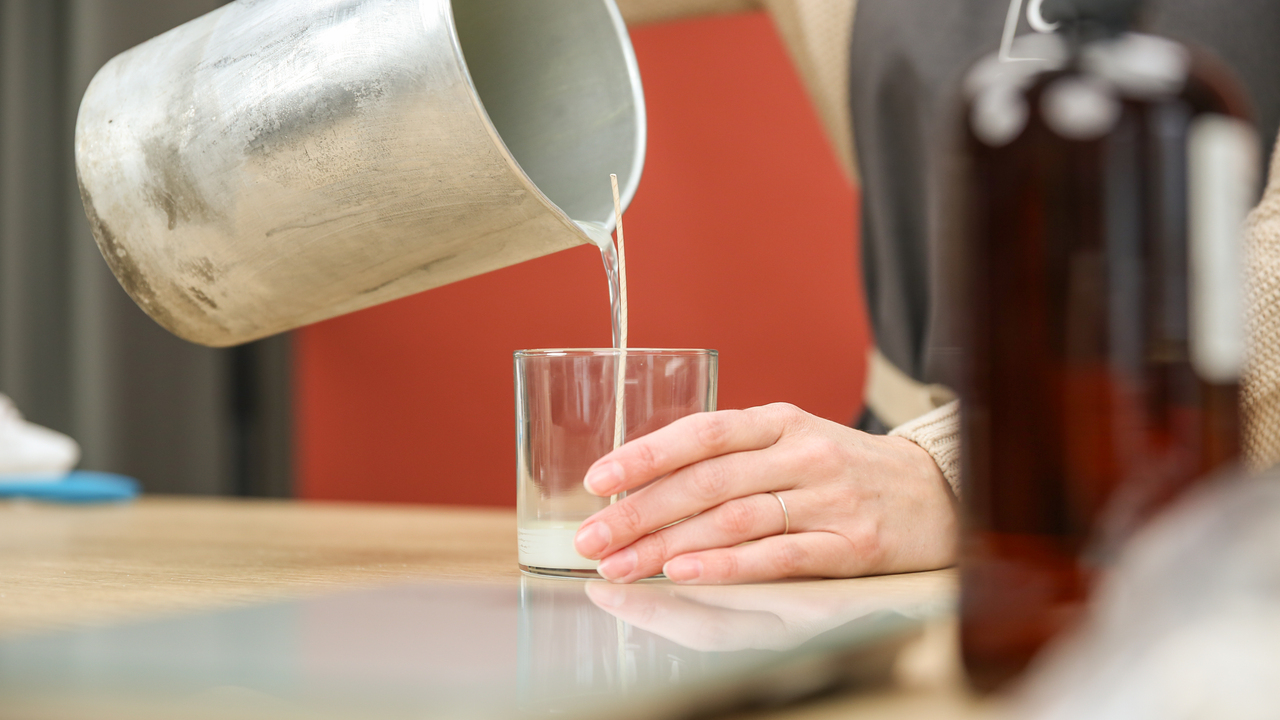 A pair of light-skinned hands holding a metal pouring pitcher dispenses melted candle wax into a clear glass jar